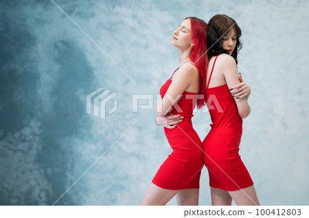 Portrait of two women dressed in identical red dresses and standing back to back. Lesbian intimacy. Portrait of two women dressed in identical red dresses and standing back to back. Lesbian intimacy. 100412803