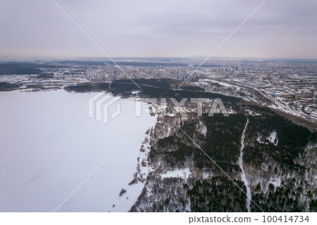 Snow-covered forest on lake shore with ice at sunset and the city on horizon, auerial view Snow-covered forest on lake shore with ice at sunset and the city on horizon, auerial view 100414734