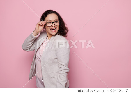 Charming dark-haired middle-aged woman smiling a toothy smile, looking aside, wearing stylish trendy eyeglasses and elegant light gray suit, isolated on pink background with free advertising space Charming dark-haired middle-aged woman smiling a toothy smile, looking aside, wearing stylish trendy eyeglasses and elegant light gray suit, isolated on pink background with free advertising space 100418937