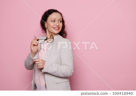 Confident middle-aged beautiful woman in formal suit, pleasant teacher or business person holding trendy glasses, smiling broadly, looking aside at copy advertising space on isolated pink background Confident middle-aged beautiful woman in formal suit, pleasant teacher or business person holding trendy glasses, smiling broadly, looking aside at copy advertising space on isolated pink background 100418940