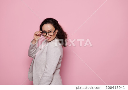 Waist-up portrait of confident successful woman in gray formal suit, looking at camera through her stylish eyeglasses, isolated on pink color background. Copy advertising space. Fashion. Style. Women Waist-up portrait of confident successful woman in gray formal suit, looking at camera through her stylish eyeglasses, isolated on pink color background. Copy advertising space. Fashion. Style. Women 100418941