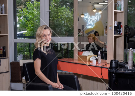 Woman with blonde wavy hair sits smiling in beauty salon 100420416