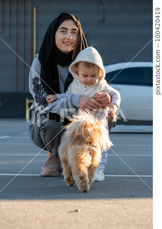 Woman in hijab enjoys walking with excited toddler and dog 100420419