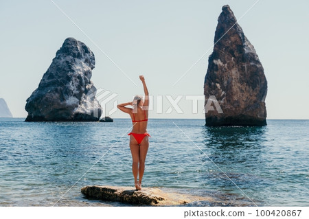 Woman summer travel sea. Happy tourist enjoy taking picture outdoors for memories. Woman traveler posing on the beach at sea surrounded by volcanic mountains, sharing travel adventure journey 100420867