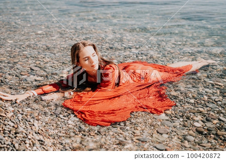 Woman travel sea. Happy tourist in red dress enjoy taking picture outdoors for memories. Woman traveler posing in sea beach, surrounded by volcanic mountains, sharing travel adventure journey 100420872