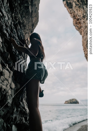 Sports Woman Climbing The Rock. Young woman With slim fit body climbing in volcanic basalt cave with beautiful sea view. The athlete girl trains in nature. Woman overcomes difficult climbing route. 100420923