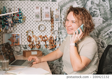 Smiling concentrated business woman talking smartphone, working on laptop in workshop. Home office. Baby accessories Smiling concentrated business woman talking smartphone, working on laptop in workshop. Home office. Baby accessories 100421132