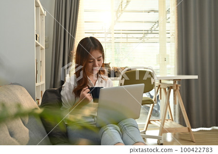 Young asian woman freelancer sitting on couch surfing internet, checking email on laptop in the morning Young asian woman freelancer sitting on couch surfing internet, checking email on laptop in the morning 100422783