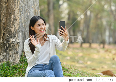 Pretty young asian woman making video call with her friends on mobile phone while sitting under a tree in public park Pretty young asian woman making video call with her friends on mobile phone while sitting under a tree in public park 100422835