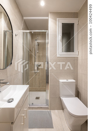 Modern compact beige tiled bathroom with glass walk-in shower, sink and mirror and white toilet with window. Concept of a tiny bathroom in a hotel or small apartment 100424499