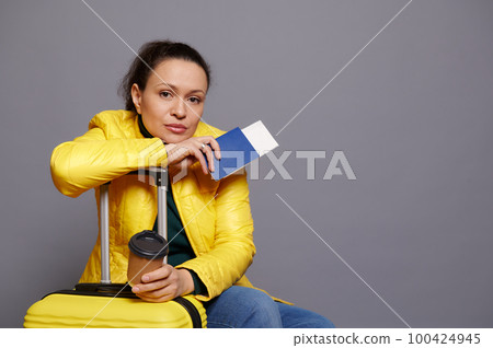 Confident beautiful Latin American woman passenger, dressed in yellow jacket, looking at camera, posing with suitcase, boarding pass and takeaway coffee in disposable cup, on gray isolated background Confident beautiful Latin American woman passenger, dressed in yellow jacket, looking at camera, posing with suitcase, boarding pass and takeaway coffee in disposable cup, on gray isolated background 100424945