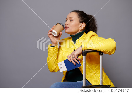 Charming pensive woman in yellow jacket, drinking coffee from disposable paper cup, sitting on a suitcase, waiting to board the flight, isolated on gray background. People, travel and tourism concept. Charming pensive woman in yellow jacket, drinking coffee from disposable paper cup, sitting on a suitcase, waiting to board the flight, isolated on gray background. People, travel and tourism concept. 100424947