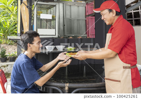 An Asian man with a hat and apron serves serabi to customers An Asian man with a hat and apron serves serabi to customers 100425261