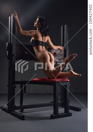 Bottomless woman sitting on BDSM bench in studio 100427405