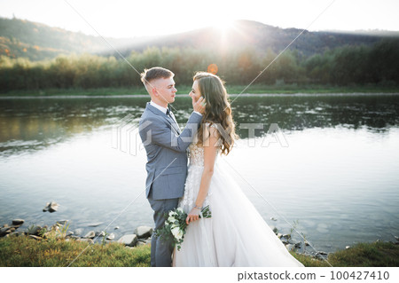 Elegant stylish happy blonde bride and gorgeous groom on the background of a beautiful river in the mountains Elegant stylish happy blonde bride and gorgeous groom on the background of a beautiful river in the mountains 100427410