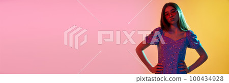 Portrait of grimacing girl with long hair wearing sunglasses keeping hands on belt zone over pink yellow background in neon light. 100434928