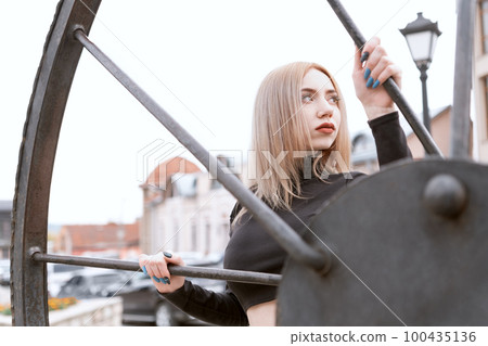 Young blonde woman girl in black turtleneck near metal wheel on urban architecture background. Young blonde woman girl in black turtleneck near metal wheel on urban architecture background. 100435136