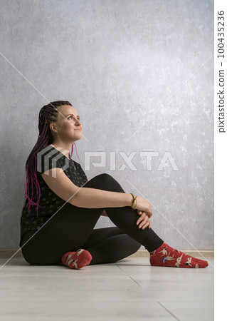 Portrait of young woman with dreadlocks sitting on the floor and thinking against gray background. Vertical frame. 100435236