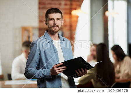 Teamleader of IT company. Young ambitious man in official clothes looking at camera in office. Blurred employees on background 100436476