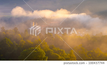 Castle Rozmberk shrouded in morning mist illuminated by the rising sun. Rozmberk nad Vltavou, Czech Republic. Aerial view from a drone Castle Rozmberk shrouded in morning mist illuminated by the rising sun. Rozmberk nad Vltavou, Czech Republic. Aerial view from a drone 100437744