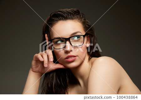 Portrait of beautiful brunette model girl with long straight hair and bare shoulders in eyeglasses over grey background. Concept of beauty, fashion, style, emotions 100438864