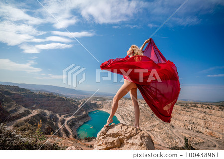 woman red dress lake mountains. Side view of a woman in a long red dress posing on a rock high above the lake. Against the background of the blue sky and the lake in the form of a heart 100438961
