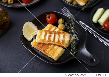 Grilled halloumi cheese in a black pan with olives, tomatoes, cucumbers, thyme and lemon. Traditional cheese of the Eastern Mediterranean. Black background, selective focus, close-up. 100439311