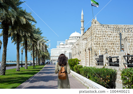 A woman in a long dress with a backpack walks along the Al Majaz embankment, Lake Khaled, Sharjah emirate. Rear view of a woman walking in the park 100440021