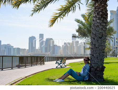 Happy tourist relaxing on lawn under palm tree in Sharjah while looking at camera. Vacation and sightseeing concept. Happy tourist relaxing on lawn under palm tree in Sharjah while looking at camera. Vacation and sightseeing concept. 100440025