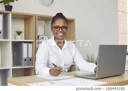 Portrait of happy African American businesswoman at office desk with paperwork and laptop 100443280