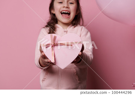 Details on cute pink heart shaped gift box with happy surprise inside it, in hands of blurred little child girl, expresshing happiness and joy, smiling looking at camera over isolated pink background 100444296