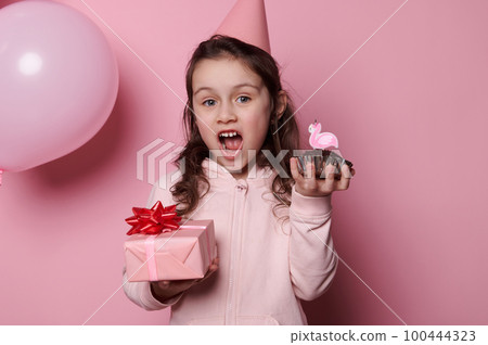 Adorable Caucasian little child girl in pink festive hat, posing with gift box and cupcake on isolated color background, expressing amazement looking at camera, celebrating her fifth birthday party Adorable Caucasian little child girl in pink festive hat, posing with gift box and cupcake on isolated color background, expressing amazement looking at camera, celebrating her fifth birthday party 100444323