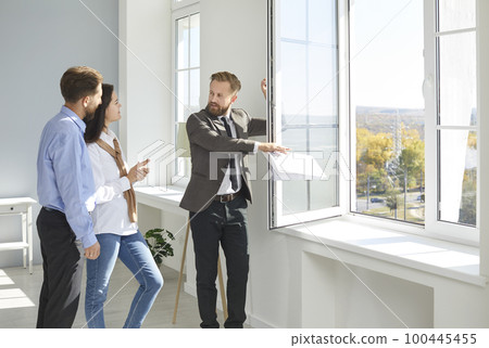 Male realtor shows apartment for sale and its view from window to young married couple. Male realtor shows apartment for sale and its view from window to young married couple. 100445455