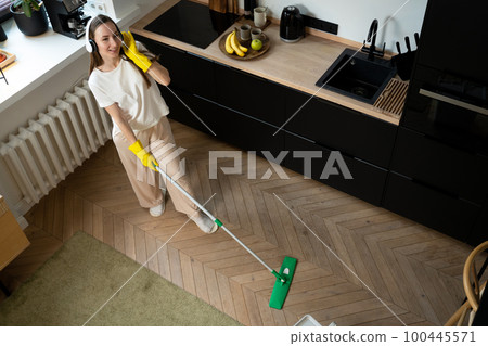 Happy beautiful young woman, housewife in rubber gloves, and white headphones, singing merrily while cleaning the kitchen floor 100445571