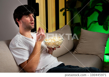Young man sitting on the couch watching an interesting movie on the TV screen, eating popcorn 100445971