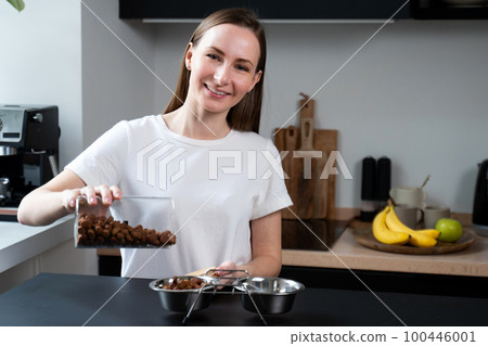 Woman pours dog food for pets into a steel bowl in the kitchen 100446001