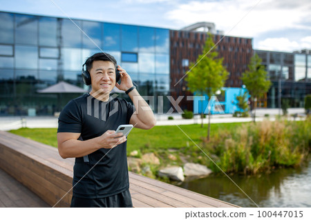 A young Asian sportsman man is sitting resting on a wooden bench near the lake and using a mobile phone, holding headphones. Smiling at the camera. A young Asian sportsman man is sitting resting on a wooden bench near the lake and using a mobile phone, holding headphones. Smiling at the camera. 100447015