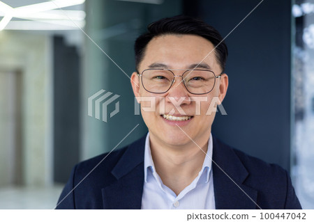 Closeup portrait successful Asian man in glasses and business jacket smiling and looking at camera adult mature businessman working inside office financier investor happy with work and achievement Closeup portrait successful Asian man in glasses and business jacket smiling and looking at camera adult mature businessman working inside office financier investor happy with work and achievement 100447042