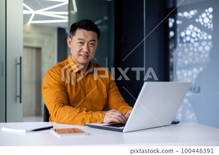 Portrait of happy and satisfied asian programmer inside office working with laptop, male freelancer in yellow shirt sitting at desk smiling and looking at camera, programming coding software. Portrait of happy and satisfied asian programmer inside office working with laptop, male freelancer in yellow shirt sitting at desk smiling and looking at camera, programming coding software. 100448391