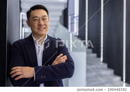 Portrait of successful asian businessman inside office building, man with crossed arms smiling and looking at camera, boss in business suit and glasses standing in hall corridor of office center. 100448392