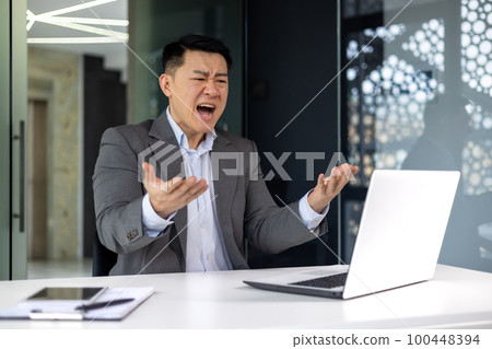 Angry boss working at work, senior experienced businessman in business suit shouting at computer, adult Asian man inside office, sitting at workplace, dissatisfied investor financier. Angry boss working at work, senior experienced businessman in business suit shouting at computer, adult Asian man inside office, sitting at workplace, dissatisfied investor financier. 100448394