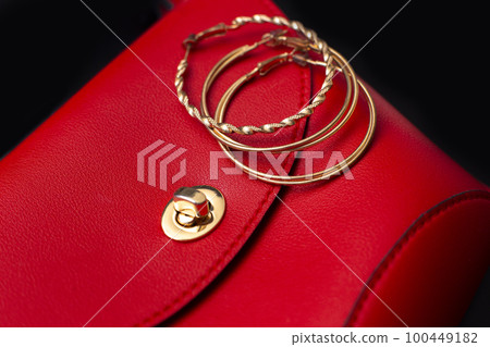 Gold ring earrings lie on a red leather bag 100449182