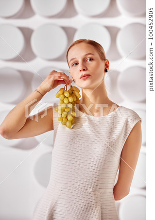 A young woman with special needs with a grape storm in her hand, a girl born without a hand, in a white dress on a white background in the rays of the sun beauty fashion concept 100452905
