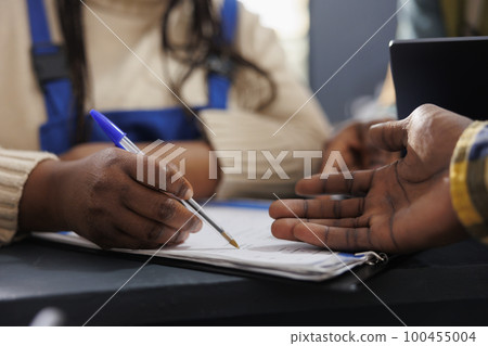 African american delivery service worker searching parcel code in clipboard. Postal warehouse employees checking goods inventory documentation and taking notes hands close up 100455004
