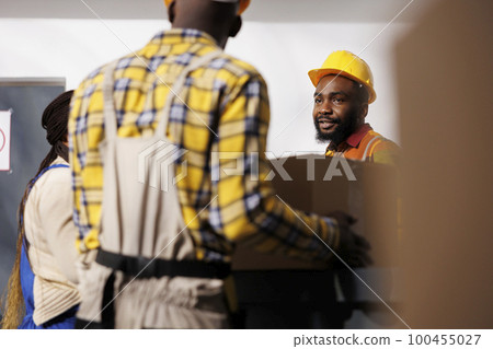 Warehouse manager looking at colleague bringing parcel to reception desk in storage room. African american storehouse employee wearing protective hard hat receiving box from package handler 100455027