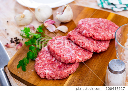 Uncooked healthy burgers on a chopping board served with heads of garlic, peppercorns, on a wooden tabletop at the restaurant 100457617