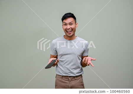 Excited Asian man holding his phone and opening his palm, standing against an isolated background Excited Asian man holding his phone and opening his palm, standing against an isolated background 100460560