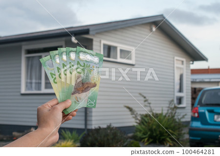 Cropped shot view of men hand holding New Zealand dollar bills in front of a single house in New Zealand. Conceptual of Real estate business. 100464281