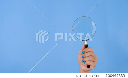 Female hands holding magnifying glass on pastel blue background in studio. Body language concept. With place for text or image, promotional content. 100469883