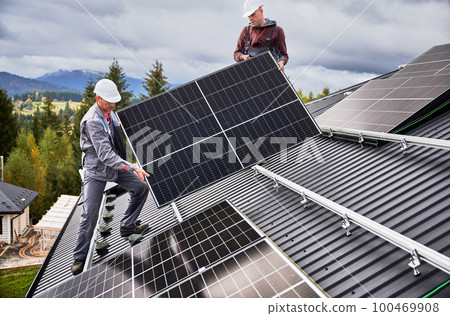 Engineers building solar panel system on roof of house. Men workers in helmets carrying photovoltaic solar module outdoors. Concept of alternative and renewable energy. 100469908